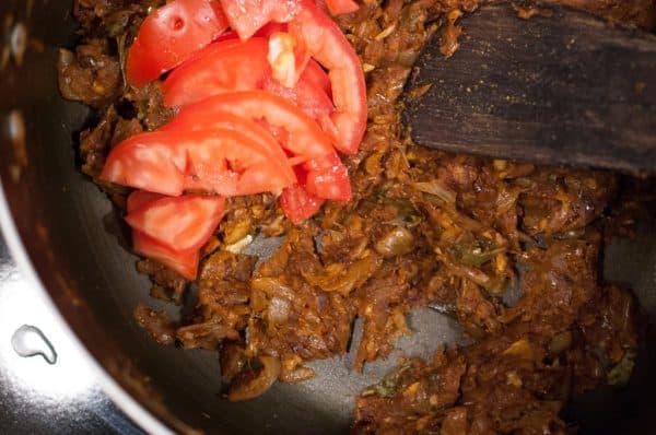 Sliced tomatoes are being added to sautéed onions and spices for Kerala Style Nadan Chicken Roast preparation.
