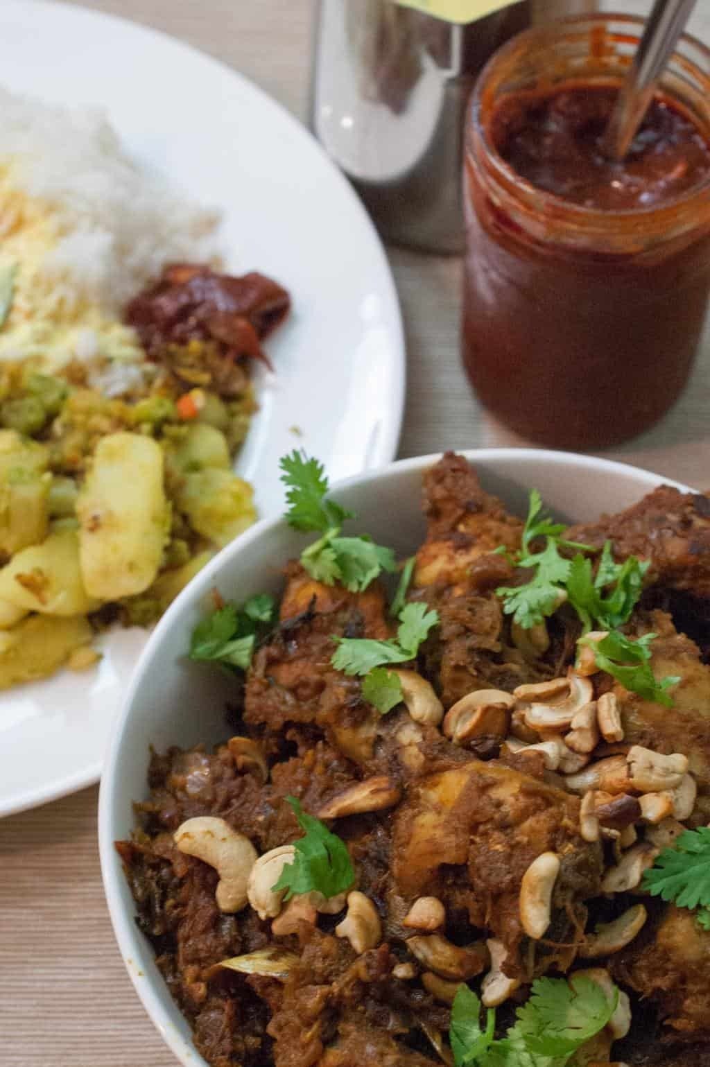 Kerala Style Chicken Roast served in a white bowl, garnished with fried cashews and coriander, alongside steamed rice, potato curry, and pickle.