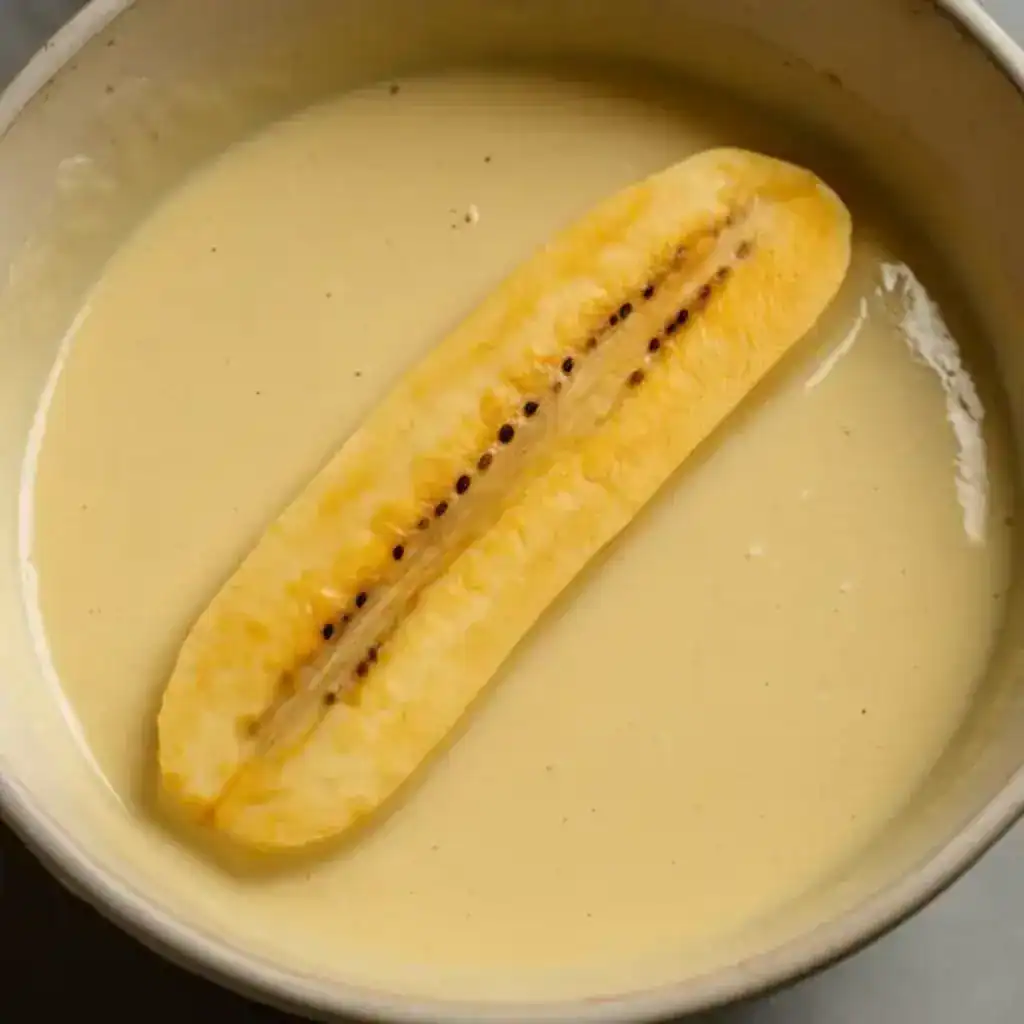 A ripe plantain slice dipped halfway into yellow batter in a ceramic bowl.