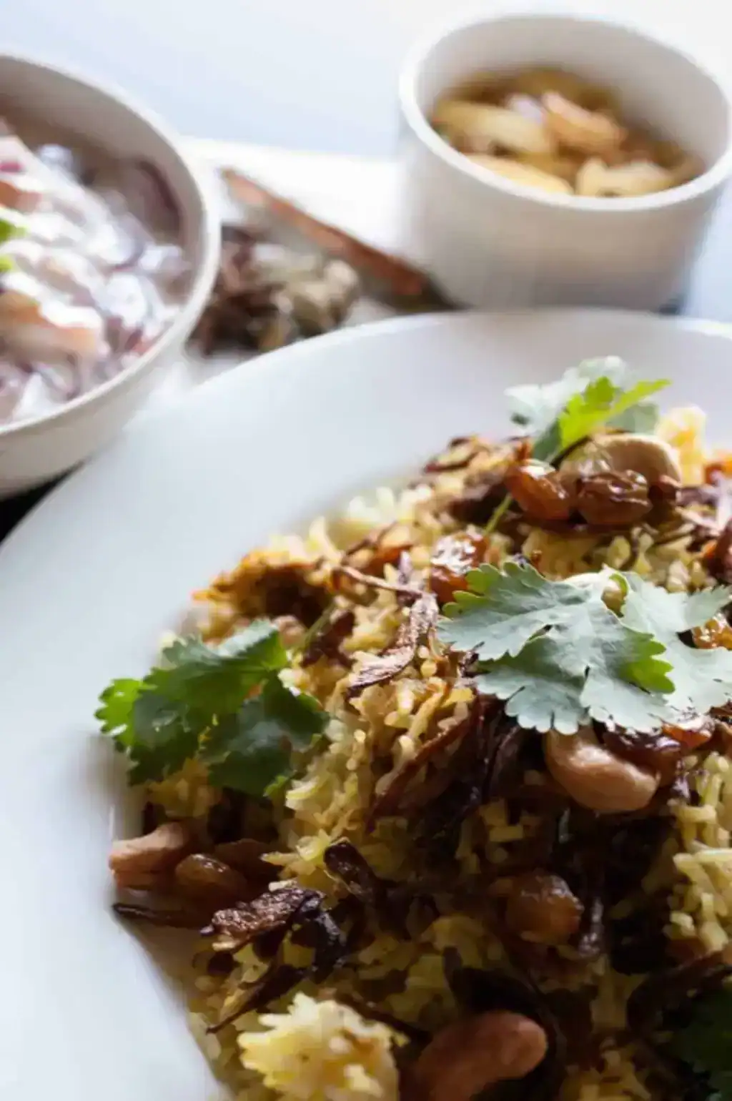 Kerala style beef/mutton biriyani served on a white plate, garnished with fried onions and cashews, with raitha on the side.