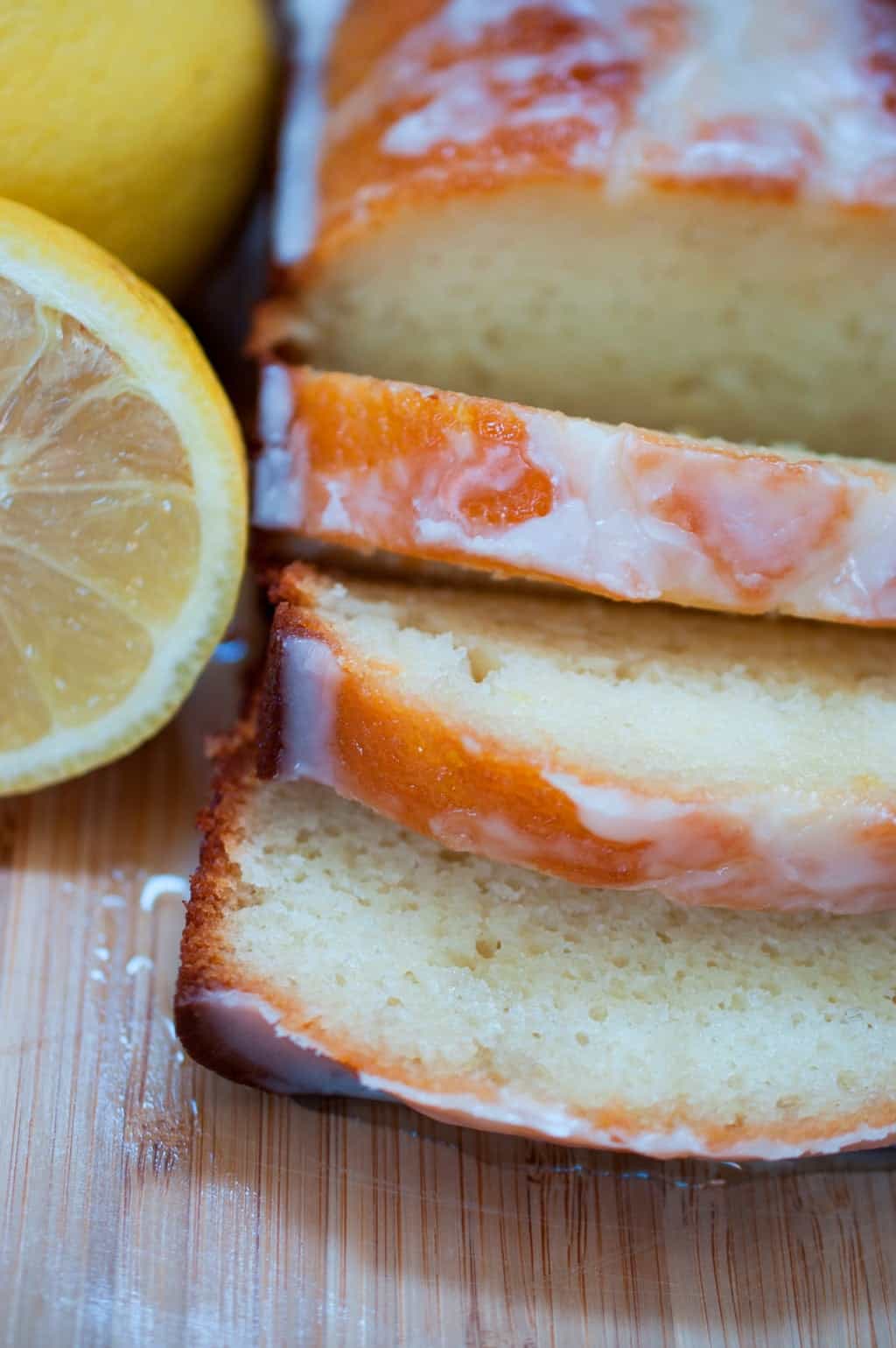 Glazed lemon yogurt cake slices with smooth texture on a wooden cutting board, with a fresh lemon beside.