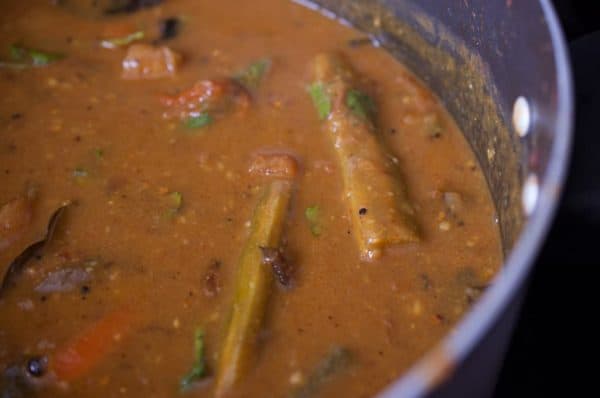 Kerala style sambar simmering in a pot with drumstick and vegetables in a reddish-brown tamarind gravy.