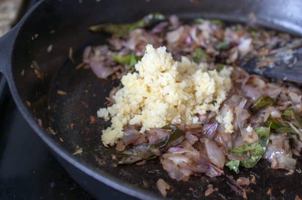 Adding ginger garlic paste to browned sautéed onions in a cast iron pan during Kerala mutton pepper fry preparation.