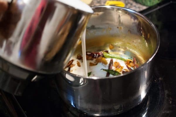 Prepared buttermilk being poured into sautéed spices and curry leaves in a pan for Kerala Moru Curry.