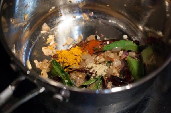 Turmeric, chilli powder, and fenugreek powder added to sautéed shallots, curry leaves, and chillies in a pan.