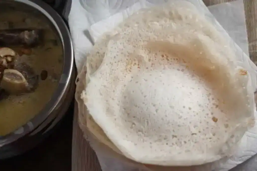 Kerala-style Appam stacked on a plate served with mutton stew in a steel bowl on the side.