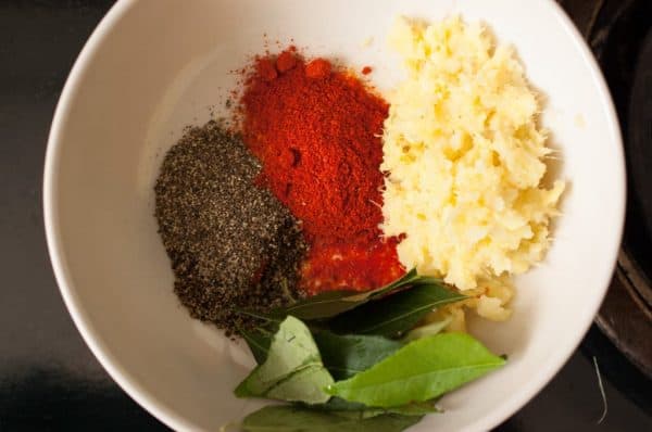 A white bowl containing red chili powder, pepper powder, curry leaves, and ginger garlic paste for Kerala-style fish fry.