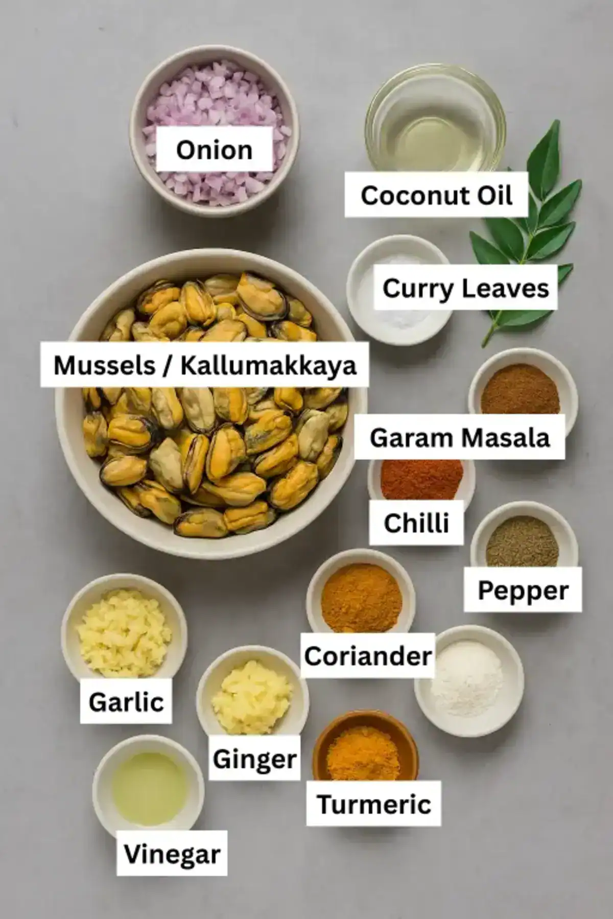Ingredients for kallumakkaya varattiyath laid out in labeled bowls on a gray background, including spices and kallumakkaya.